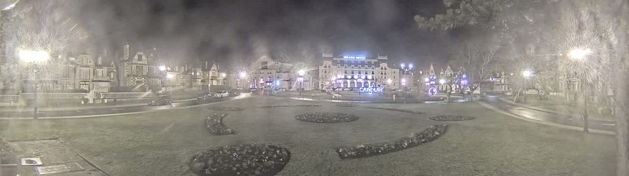 A nighttime view of a town square with buildings, streetlights, and landscaping, under hazy, snowy conditions.