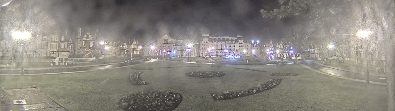 A nighttime scene shows a town square with several buildings, including a large hotel, surrounded by landscaped gardens and streetlights in snowy or hazy conditions.