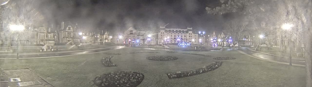A nighttime view of a town square with several buildings, including a large hotel, surrounded by landscaping and lit by streetlights in snowy conditions.