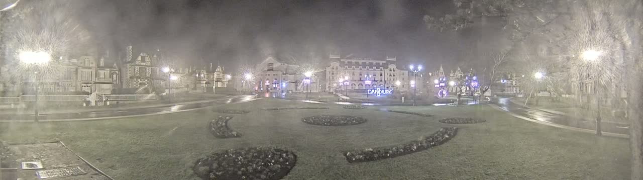 A nighttime scene of a town square with several buildings, wet pavement, and landscaping, under snowy conditions.