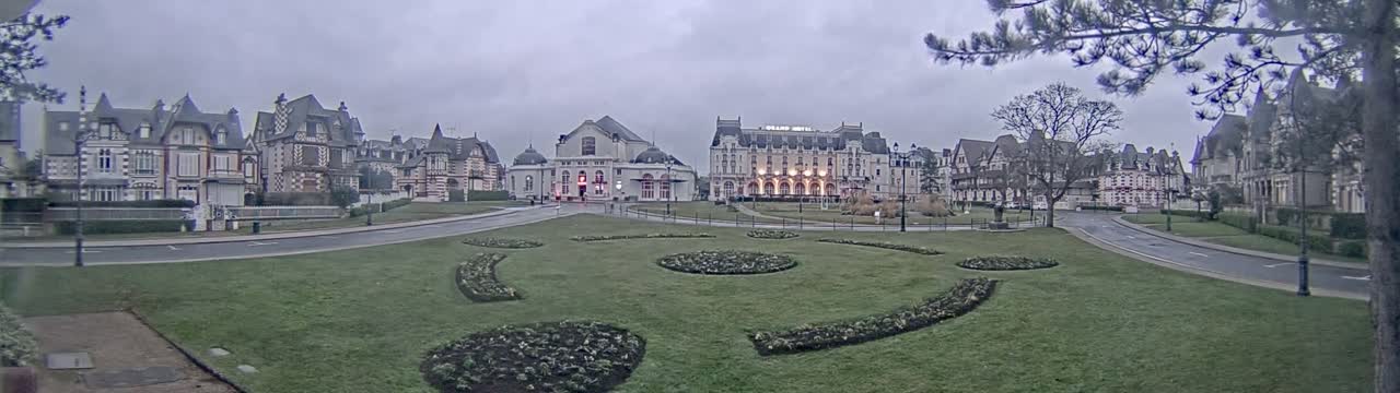 A row of ornate buildings, including a large hotel, sits across a grassy plaza with curving flower beds under an overcast sky.