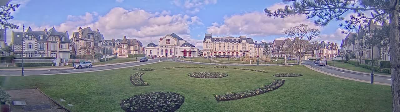 A grassy plaza with flowerbeds is surrounded by several large buildings under a partly cloudy sky.