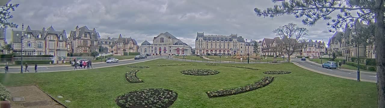 A town square with several large buildings and manicured lawns, under a cloudy sky.