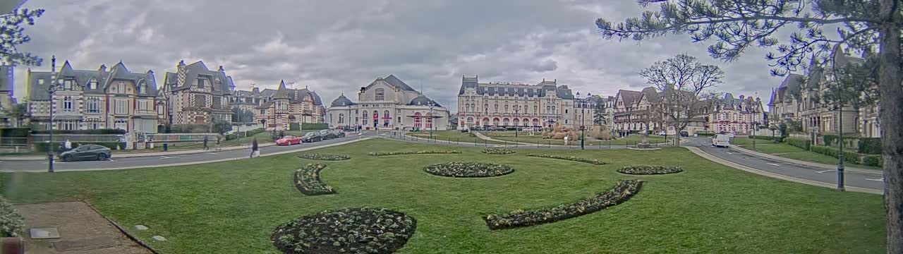 A grassy square with ornate flowerbeds is situated in front of a row of grand, multi-storied buildings under a cloudy sky.