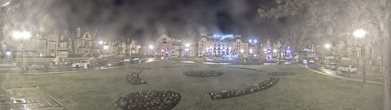 A nighttime view of a town square with several buildings, cars parked along the street, and decorative landscaping in the foreground, under rainy conditions.