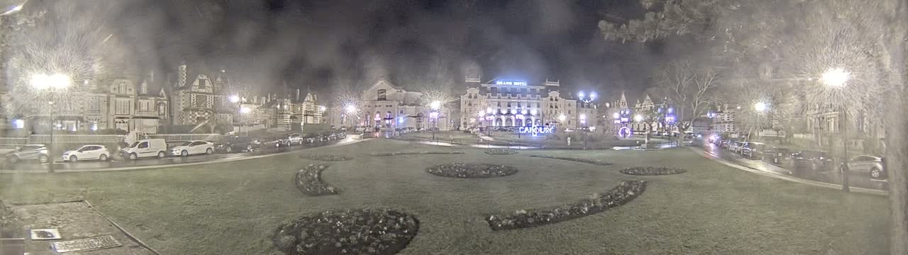 A nighttime view of a town square with several buildings, parked cars, and landscaped areas, under slightly hazy or snowy conditions.