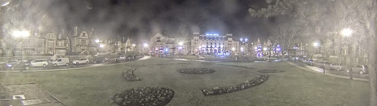 A nighttime view of a town square with several buildings, parked cars, and landscaped flower beds under hazy conditions.