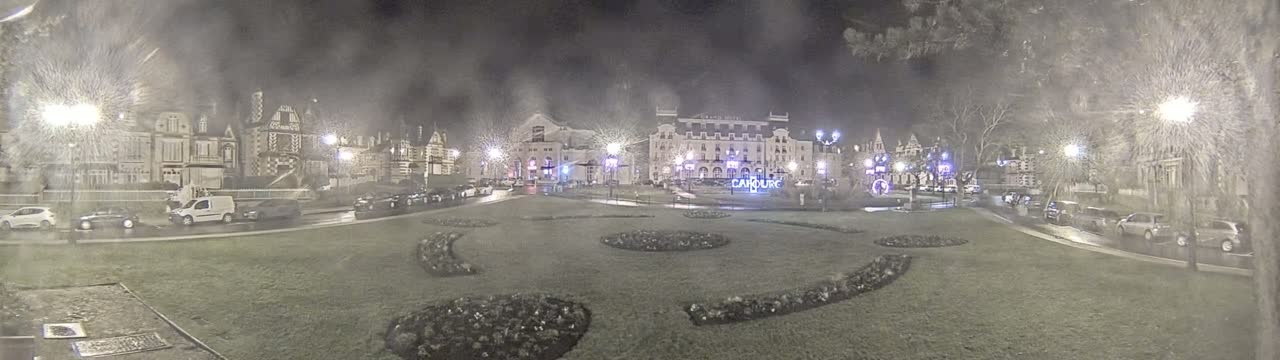 A nighttime view of a town square with several buildings, cars parked along the sides, and patterned landscaping in the center, under a hazy, possibly snowy or rainy sky.