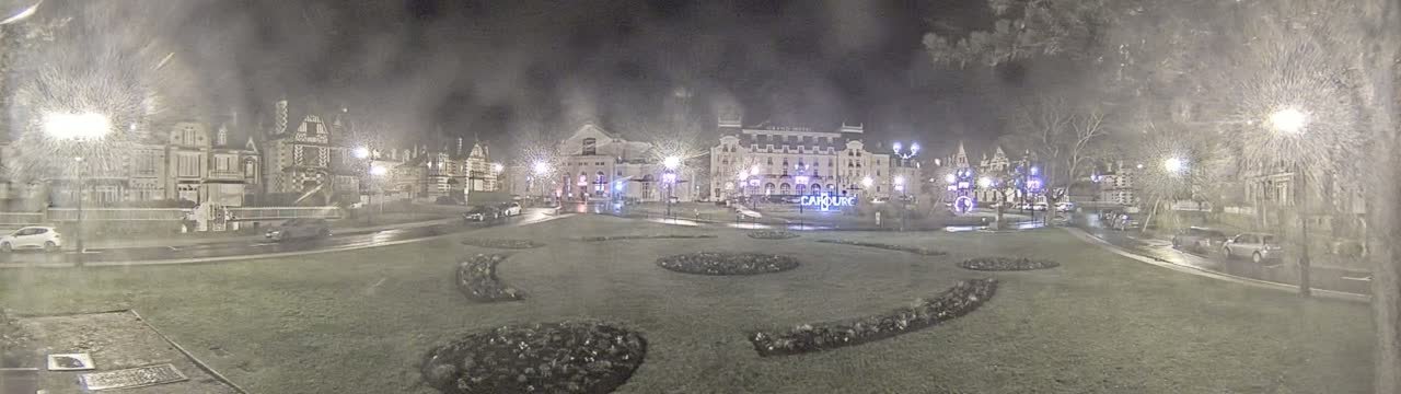 A nighttime, snowy scene shows a town square with several buildings, cars, and decorative landscaping.