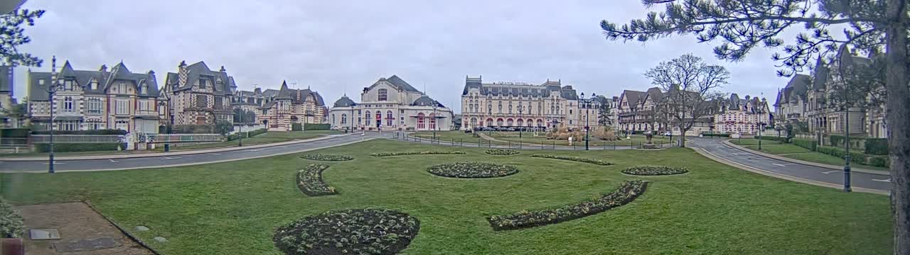 A wide, grassy expanse with ornate flower beds is situated in front of a row of large, elegant buildings under an overcast sky.