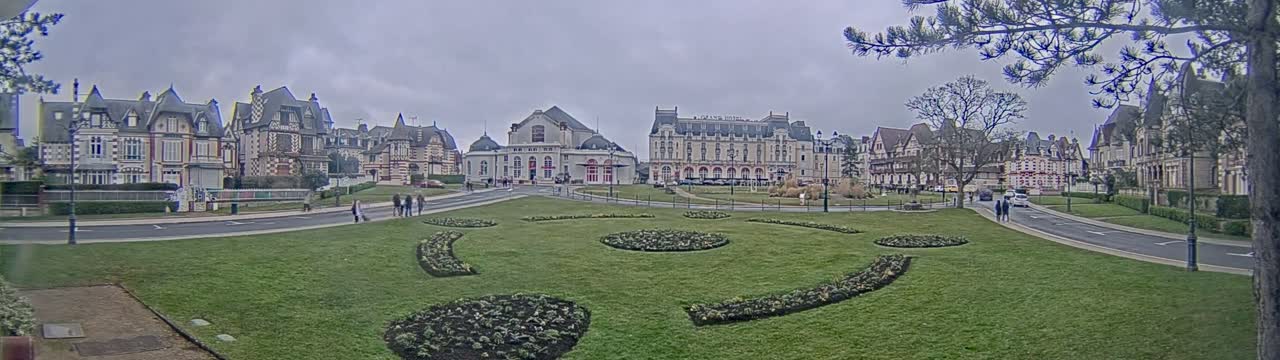 A grassy plaza with decorative flowerbeds is centered in front of a row of ornate buildings under an overcast sky.