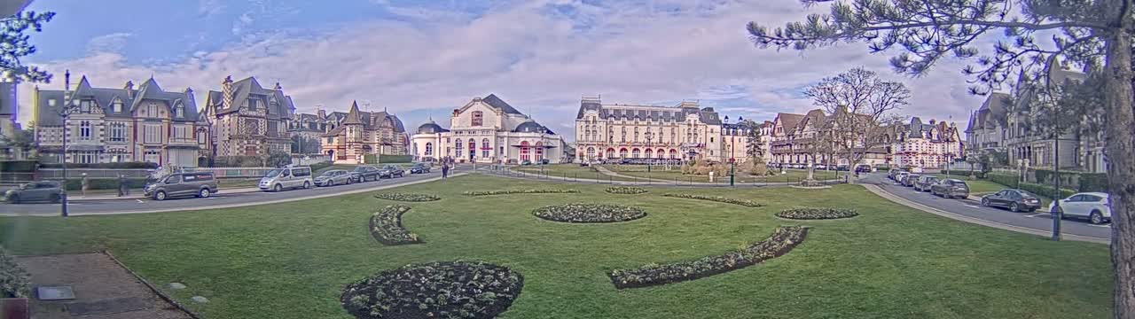A grassy plaza with landscaped flowerbeds is situated in front of several large buildings under a partly cloudy sky.