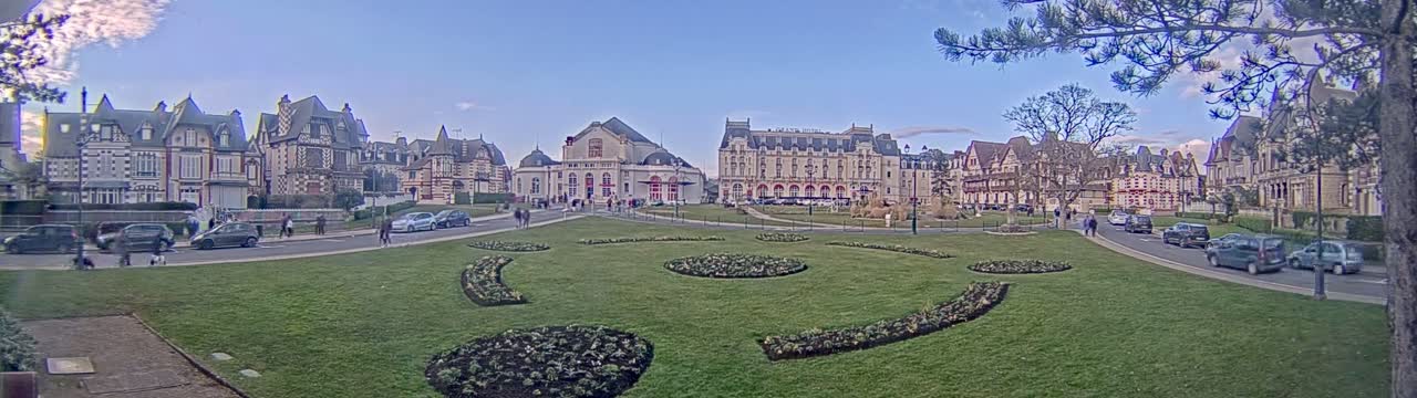 A grassy town square with decorative flowerbeds is surrounded by several large buildings, including a theater and hotels, under a mostly clear sky.
