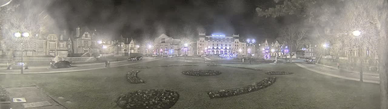 A nighttime, foggy view of a town square with several buildings, cars, and decorative landscaping.