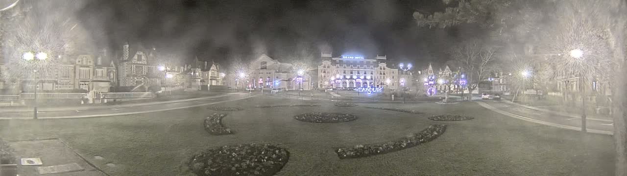 A snow-covered town square at night, illuminated by streetlights, features several buildings, including a large hotel, and landscaped flowerbeds.