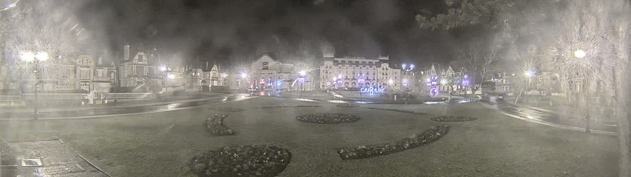 A nighttime view of a town square with several buildings, wet pavement, and decorative landscaping, under a hazy or snowy atmosphere.