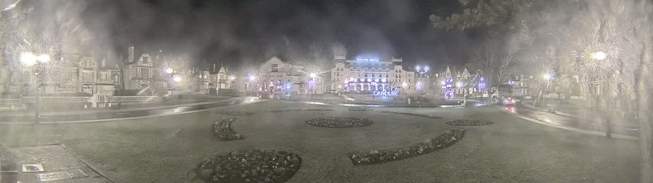 A nighttime view of a town square, possibly during a light snowfall, showing several buildings including a large hotel, decorated with lights, and landscaped gardens.