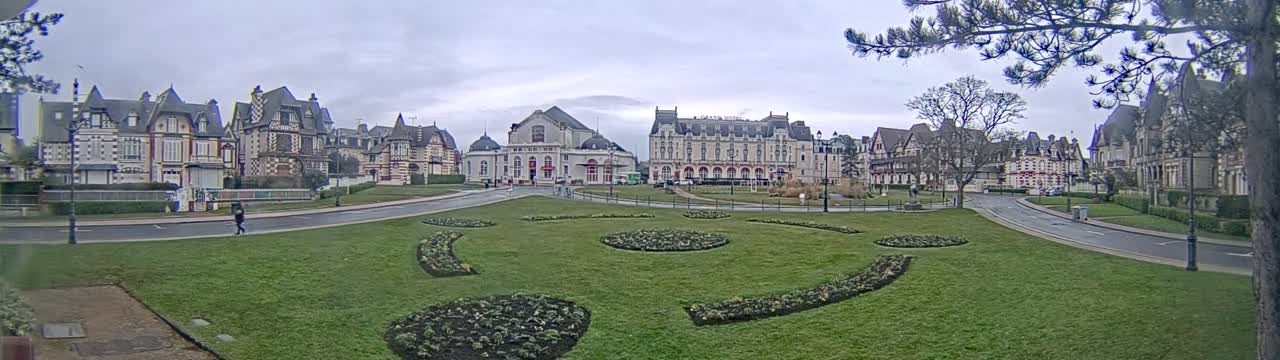 A grassy plaza with decorative flowerbeds is situated in front of a row of grand, ornate buildings under a cloudy sky.