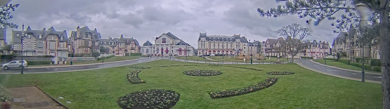 A grassy plaza with ornate flowerbeds sits in front of a row of elegant, multi-storied buildings under a cloudy sky.