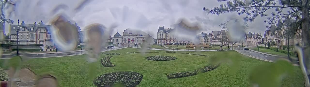 A rain-streaked view shows a grassy area with landscaped flowerbeds in front of several large, ornate buildings on a cloudy day.