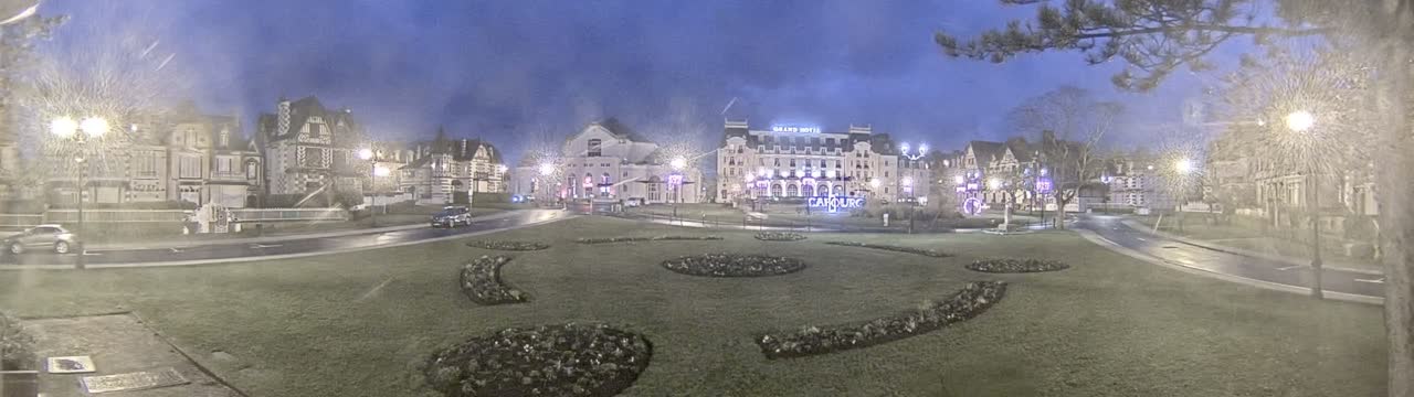 A town square at night, under a dusky sky, features several buildings illuminated by streetlights, with landscaped gardens in the foreground.