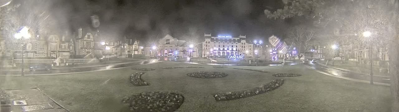 A rain-slicked town square at night, featuring several buildings, including a large hotel, and landscaped areas with flowerbeds.