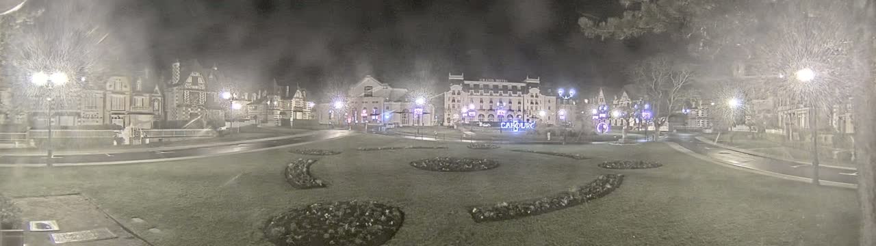 A nighttime view of a town square with several buildings, some decorated with lights, under hazy or snowy conditions.