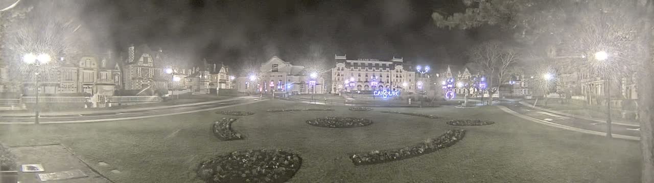 A snowy night scene shows a town square with several buildings, some lit up, and landscaped flower beds.