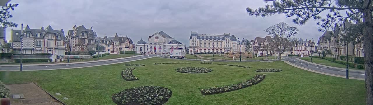 A grassy plaza with landscaped flowerbeds is surrounded by several large, ornate buildings under an overcast sky.