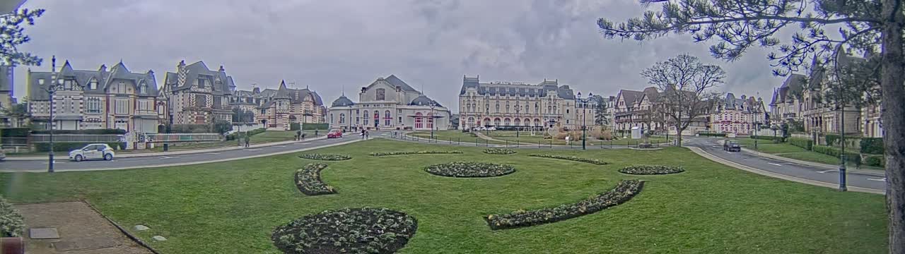 A wide, grassy park area with patterned flowerbeds is surrounded by several large, ornate buildings under a cloudy sky.