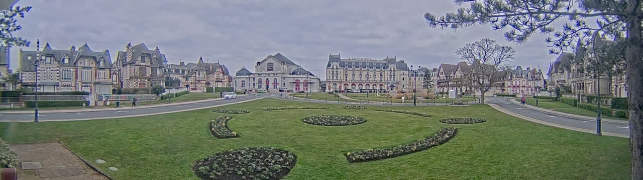 A grassy park with geometric flowerbeds is situated in front of a row of ornate European-style buildings under a cloudy sky.