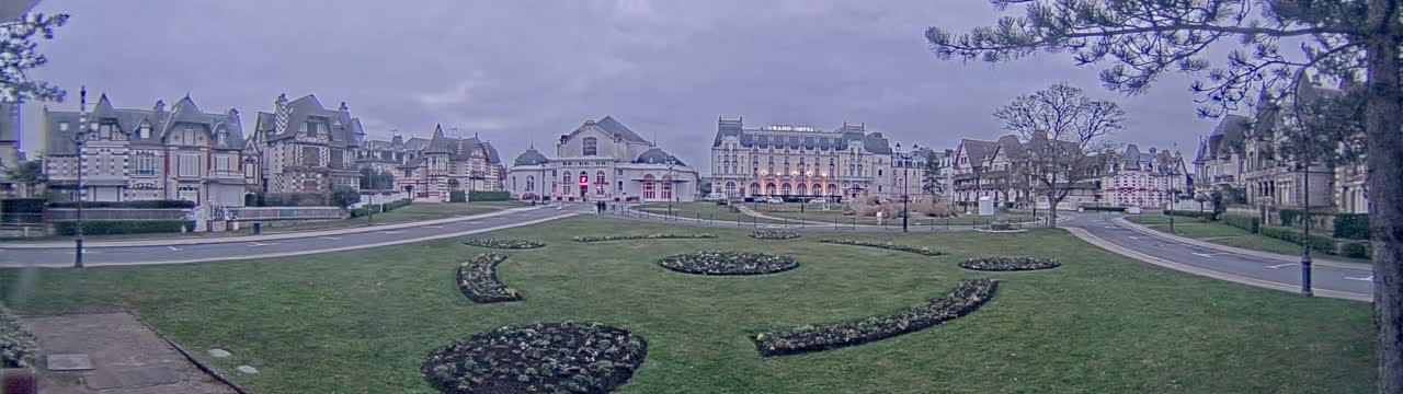 A grassy square with ornate flowerbeds sits in front of a row of large, ornate buildings on an overcast day.