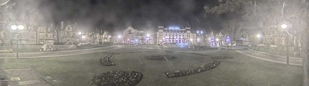 A nighttime view of a town square with several buildings, including a large hotel, surrounded by landscaping and lit by streetlights under hazy, possibly snowy conditions.