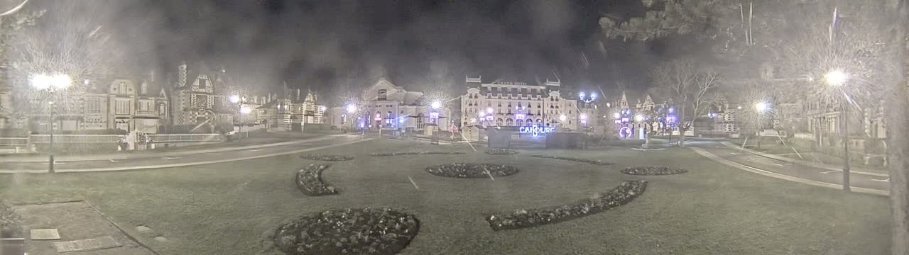 A nighttime view of a town square with several buildings, some ornate, and landscaped areas, under hazy or snowy conditions.