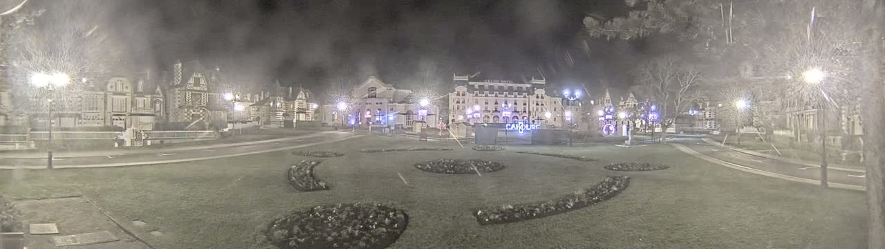 A nighttime view of a town square with several buildings, some decorative landscaping, and streetlights in snowy conditions.