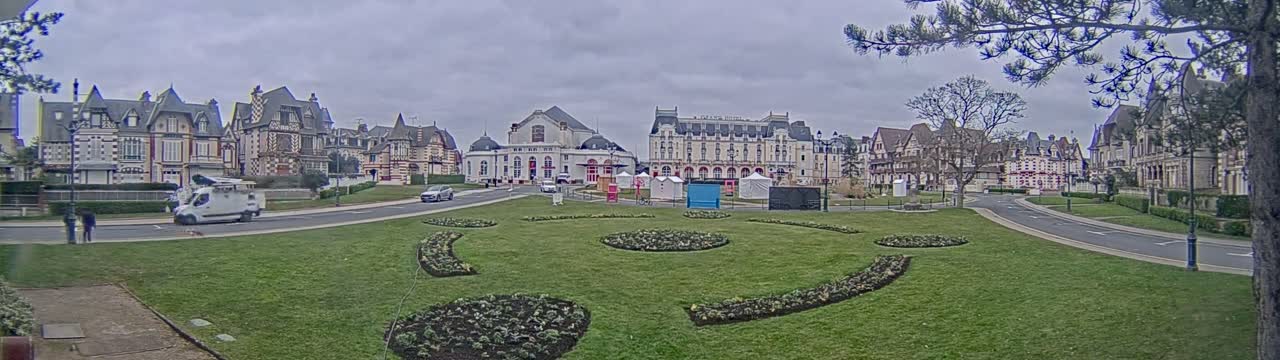 A grassy plaza with patterned flowerbeds is situated in front of a row of ornate buildings under an overcast sky.