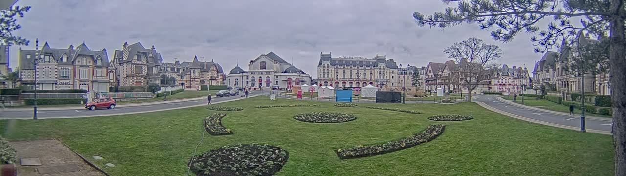 A grassy plaza with ornate flowerbeds is situated in front of several large, ornate buildings under a cloudy sky.