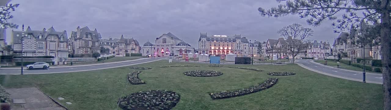 A row of large, ornate buildings sits on a grassy square with curving flowerbeds under an overcast sky.
