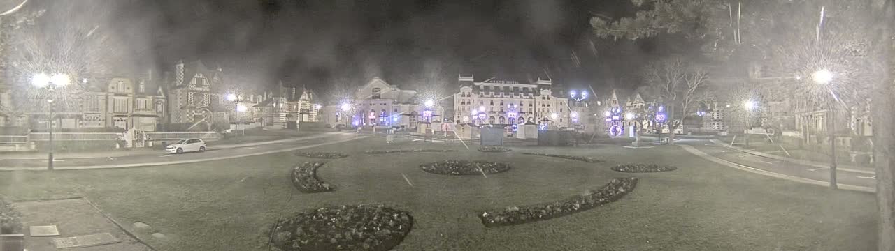 A snow-covered town square at night, illuminated by streetlights, features several buildings, including a large hotel, and landscaped areas with curved flowerbeds.