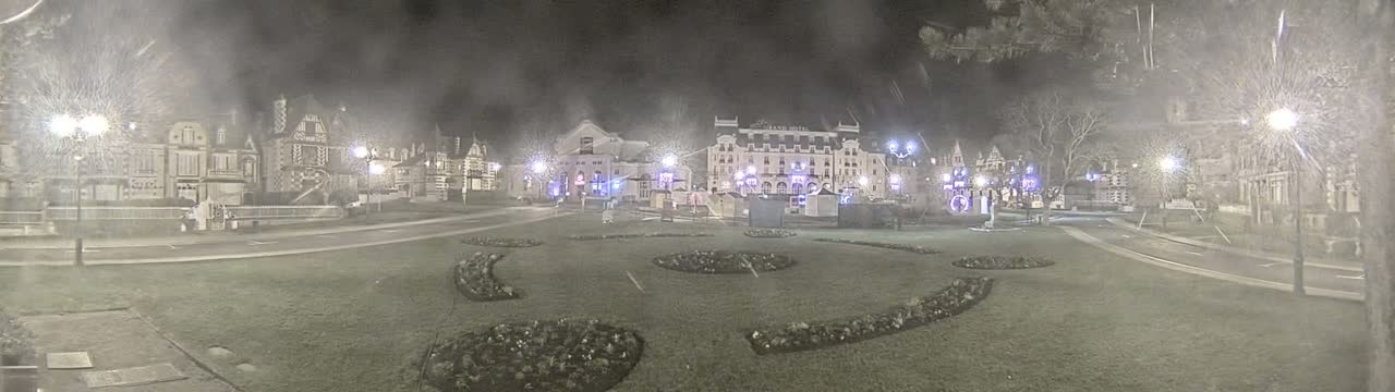 A nighttime scene of a town square with several buildings, some decorative landscaping, and lightly falling snow.