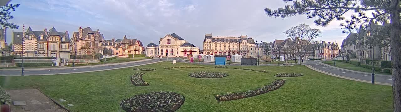 A grassy plaza with flowerbeds is in front of a row of ornate buildings under a partly cloudy sky.