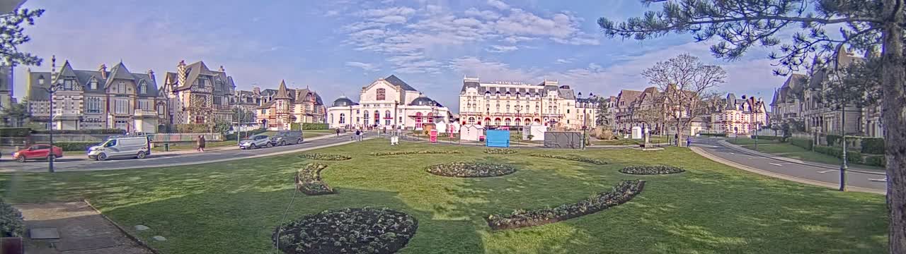 A grassy town square with manicured flowerbeds is situated in front of several ornate European-style buildings under a partly cloudy sky.