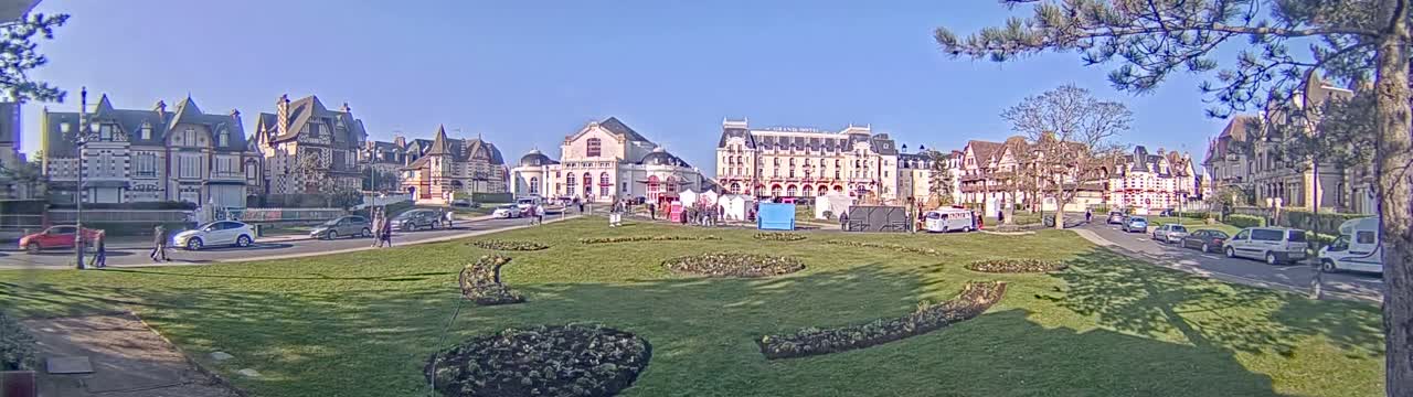 A sunny day reveals a town square with several multi-story buildings, cars parked along the roadside, and a grassy area with flowerbeds in the center.