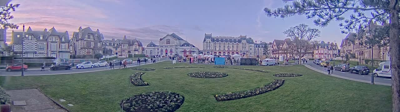 A town square with several large buildings and parked cars is surrounded by a grassy area with decorative flowerbeds under a partly cloudy twilight sky.
