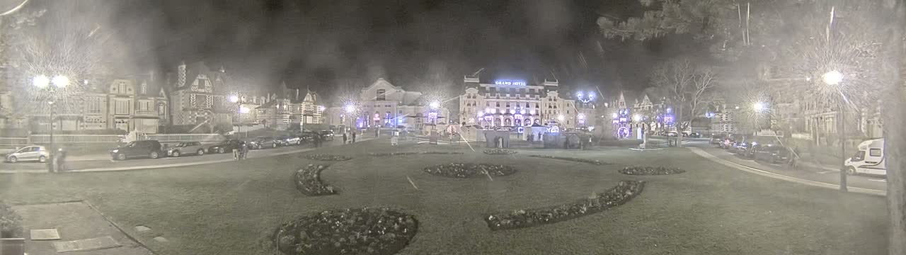 A nighttime, hazy view of a town square with parked cars, several buildings, and landscaped areas.