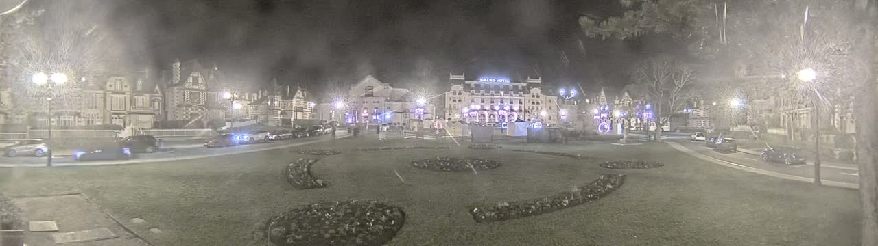 A snow-covered town square at night, featuring several buildings, cars parked along the street, and decorative landscaping.