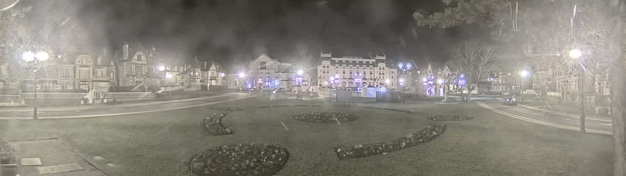 A nighttime scene shows a town square with several buildings, including a large hotel, surrounded by snow-covered ground and sparsely leafed trees under a light snowfall.