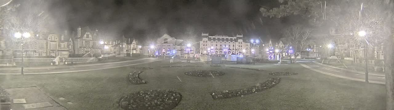 A snow-covered town square at night, illuminated by streetlights, features several buildings, including a large hotel, and landscaped areas with low-lying plantings.