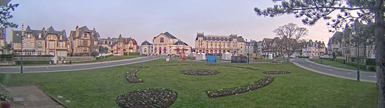 A wide, grassy square with ornate flowerbeds sits in front of a row of large, elegant buildings under a pale, dusky sky.