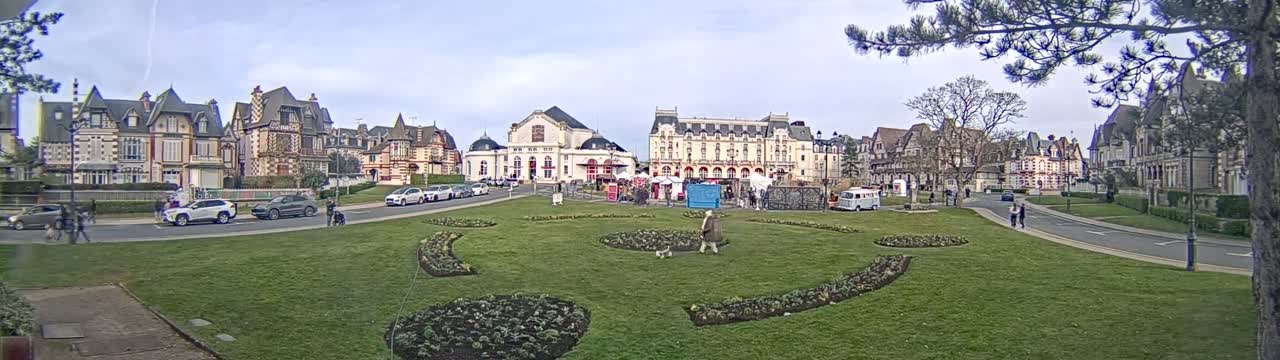 A town square with several large buildings and a grassy area with flowerbeds is visible under a partly cloudy sky.